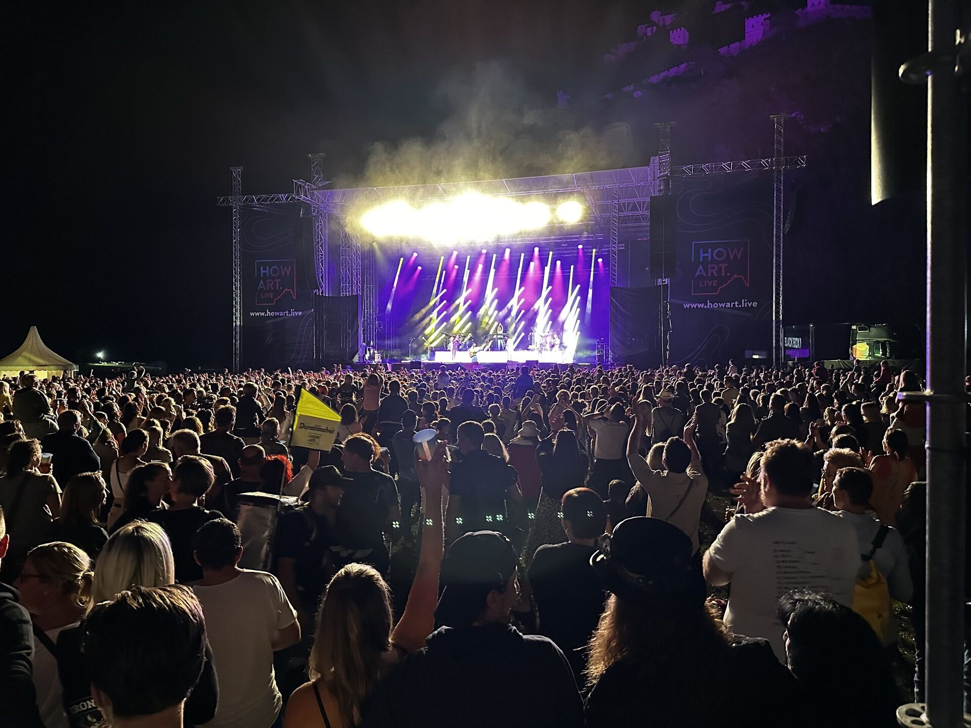 Großes Open-Air-Nachtkonzert mit hell beleuchteter Bühne, dichtes Publikum im Vordergrund, Burg auf dem Hügel dahinter