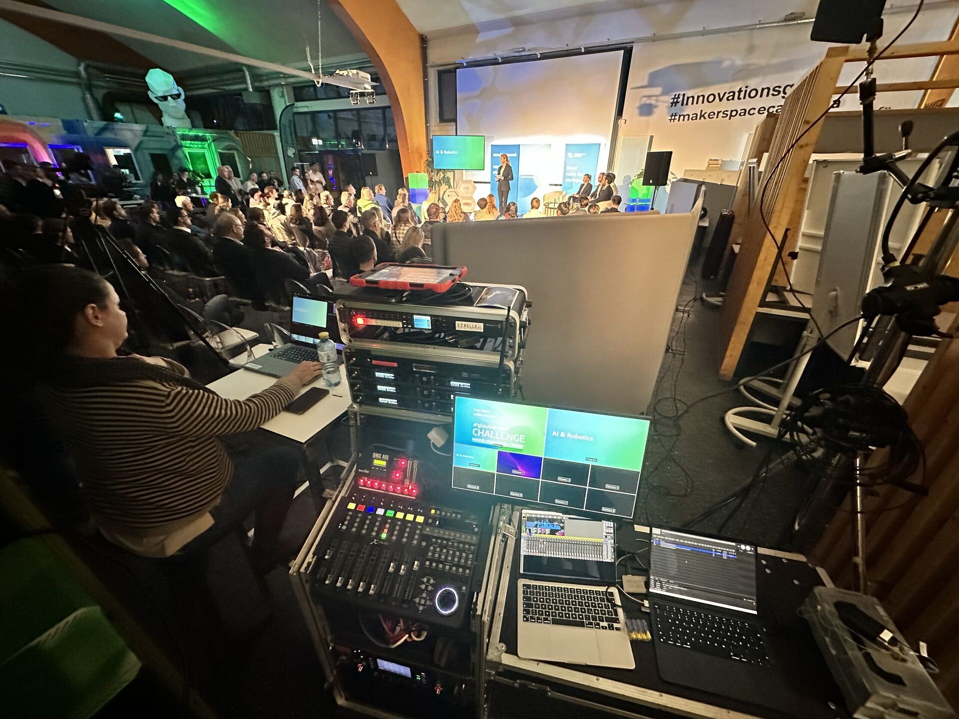 Technikerin an der Regie mit Zuspieler-Rack, Mischpult und Laptops, im Hintergrund Publikum und Bühne mit Screens