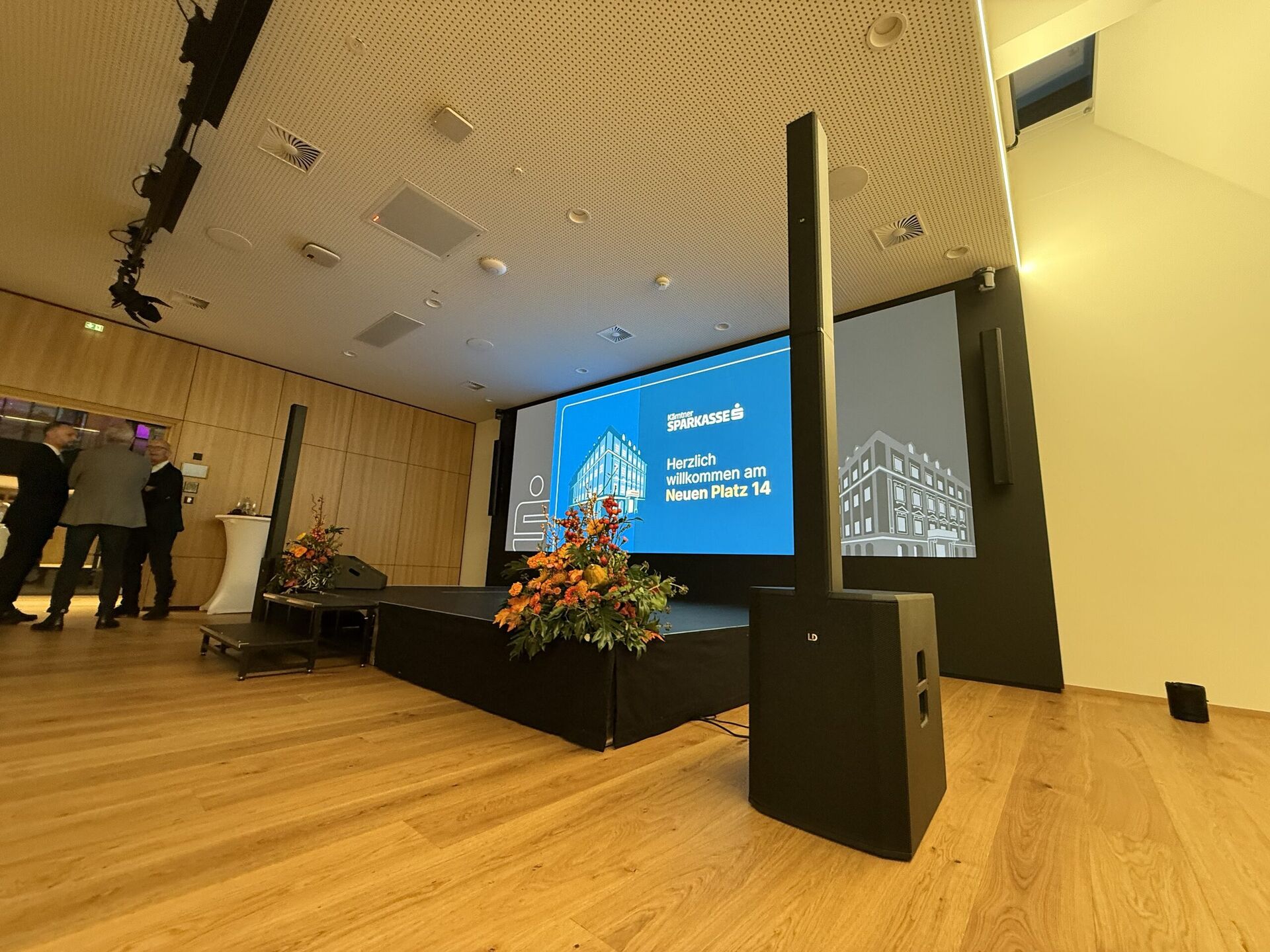 Säulen-PA-Lautsprecher neben einer kleinen Bühne in einem holzvertäfelten Veranstaltungssaal, Präsentationsscreen und Blumengesteck, Gäste im Gespräch