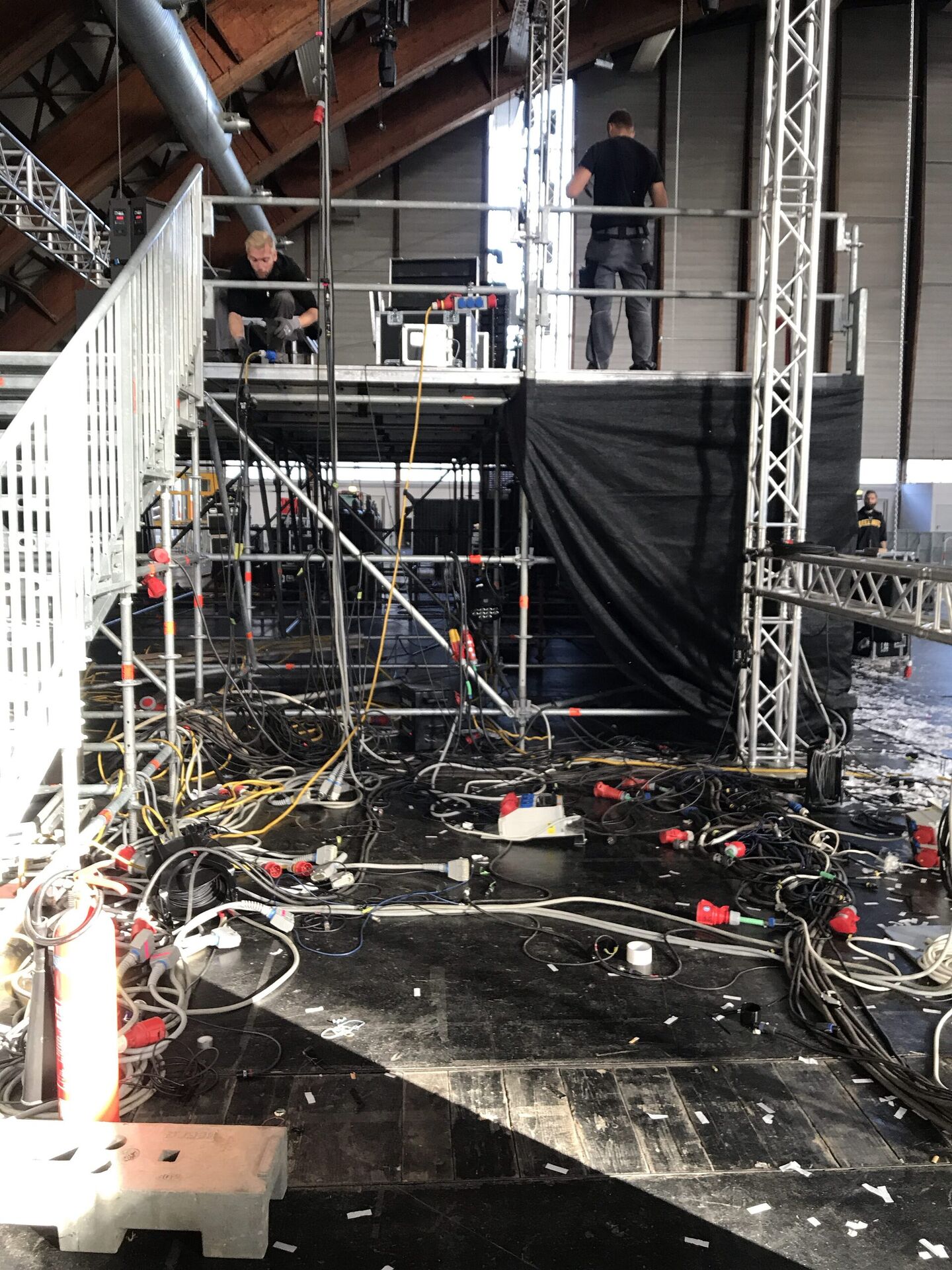 Backstage-Bereich mit vielen Stromkabeln und CEE-Steckern am Boden, Techniker an einem Rack auf dem Podest, Traversen im Hintergrund